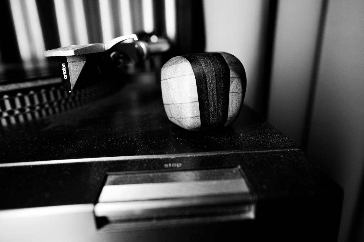 Black and white photo showing a small striped wooden sculpture on a record player turntable, with vinyl records and audio equipment in soft focus background.