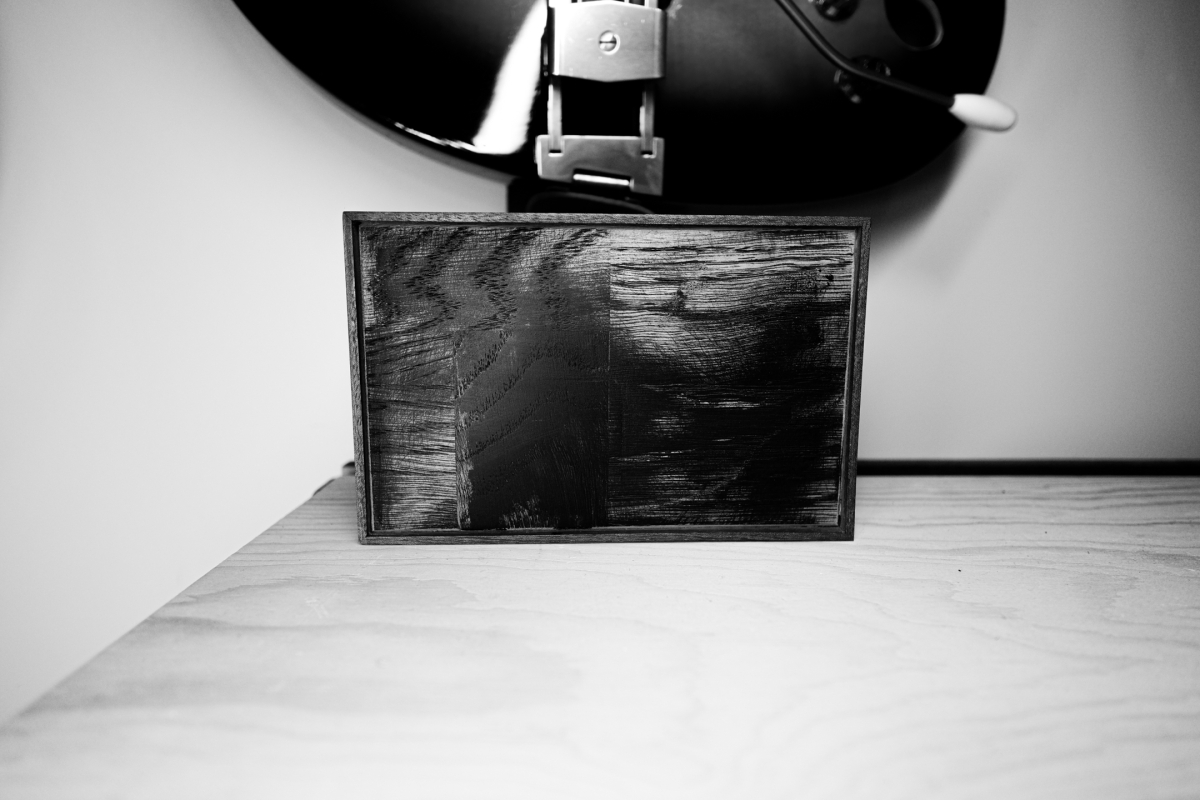 Black and white image showing a framed artwork with dark sketchy textures on a wooden surface, with a desk lamp visible above