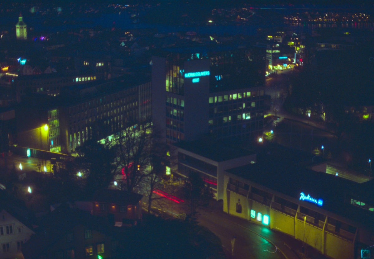 Aerial night view of a city skyline with illuminated buildings, neon signs including "Radisson," a church tower, and red light trails from traffic.