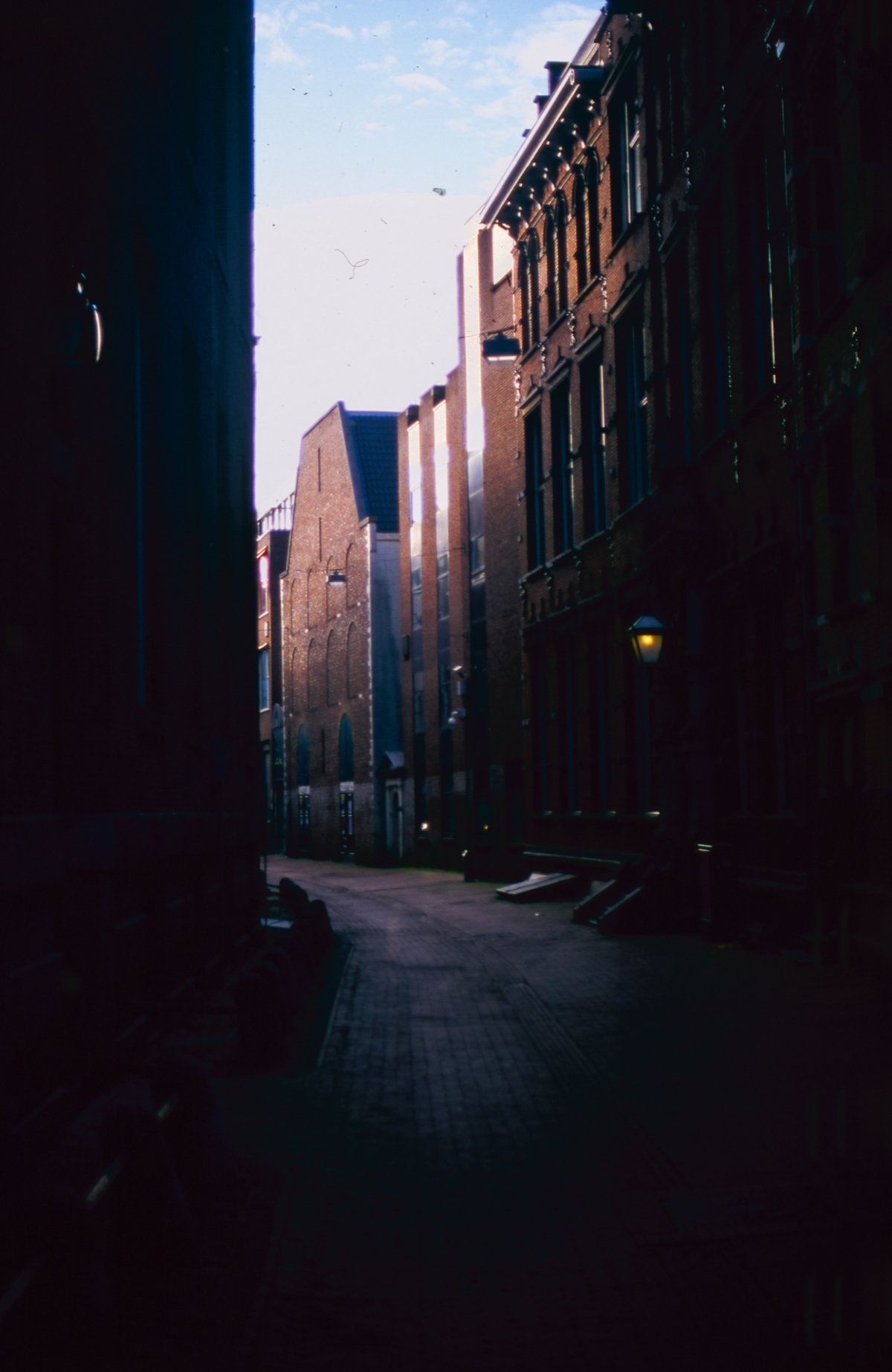 Narrow brick alleyway at dusk with warm lighting, tall red brick buildings creating shadows, moody urban atmosphere with purple-pink sky visible above.