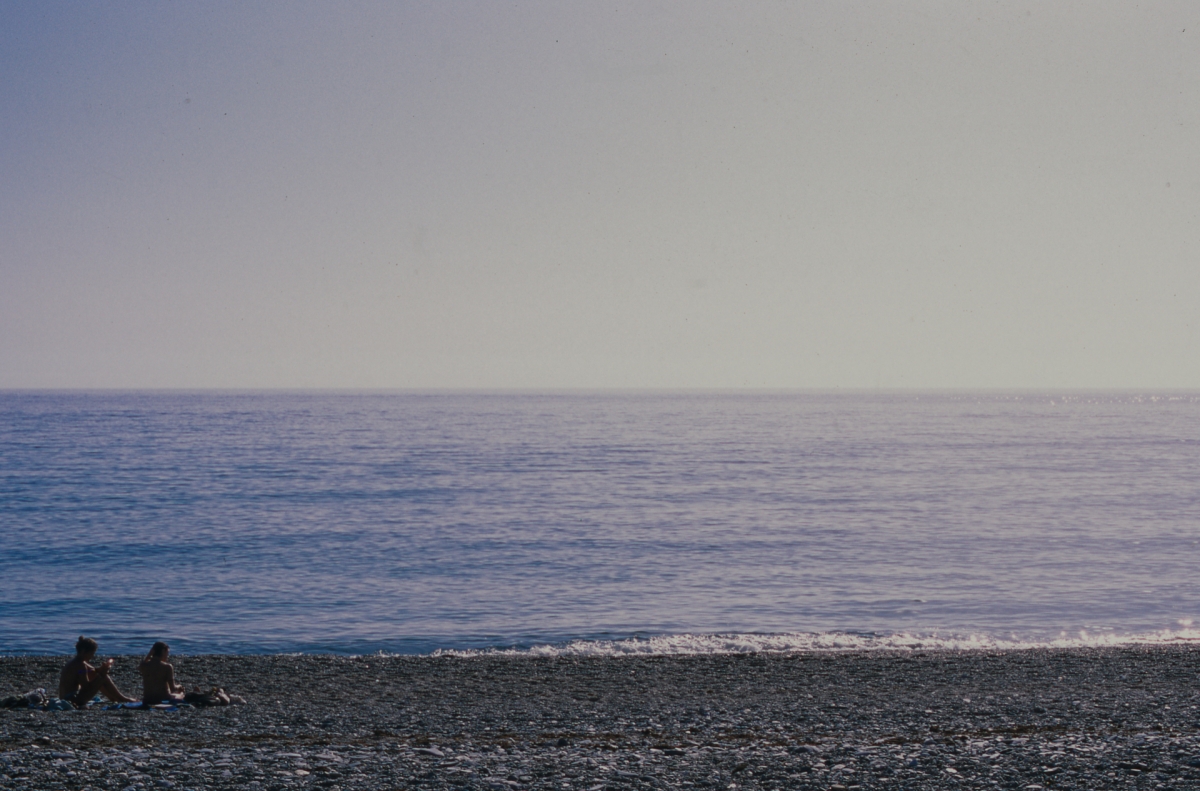 Two people sitting on rocky pebble beach beside calm blue ocean under hazy purple-gray sky at dusk or dawn