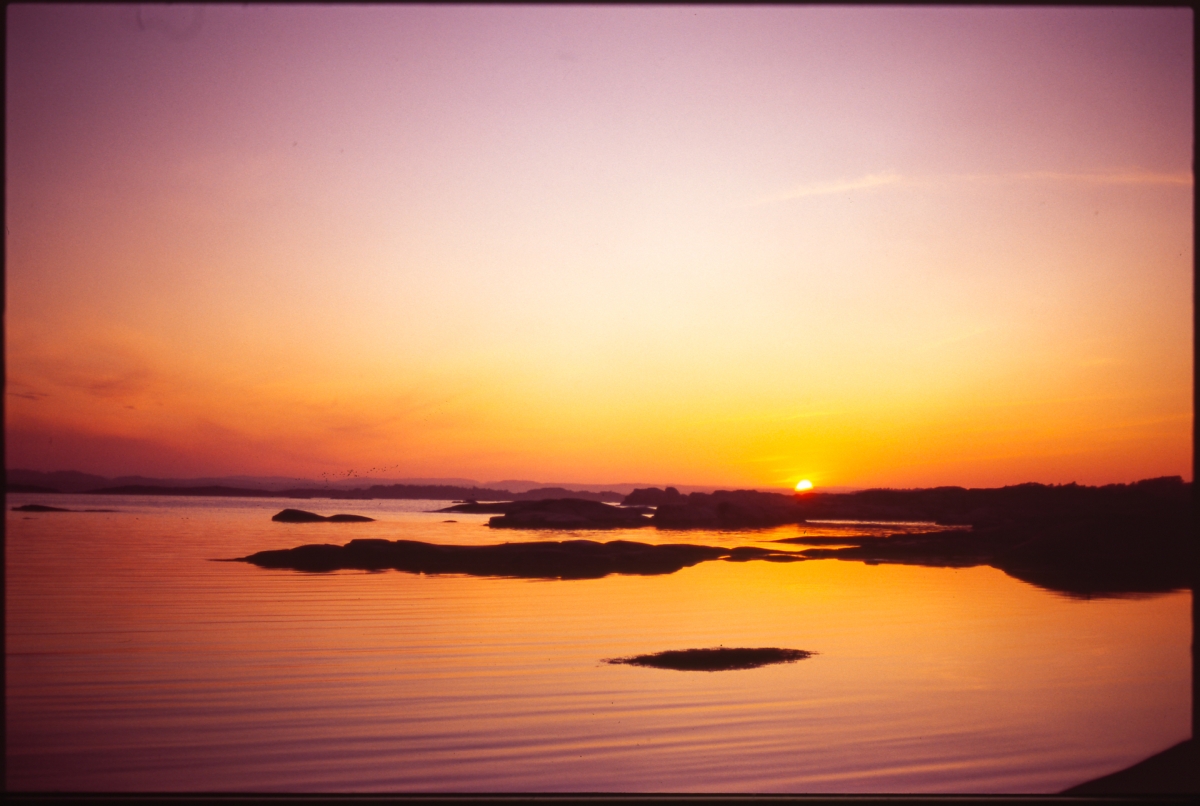 Serene sunset over calm water with silhouetted rocky islands, sky ablaze in purple, orange and yellow hues reflected on surface