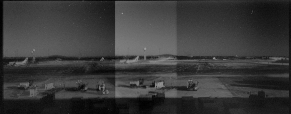 Black and white panoramic image showing three sequential frames of an open field with scattered buildings and vehicles under a gray sky