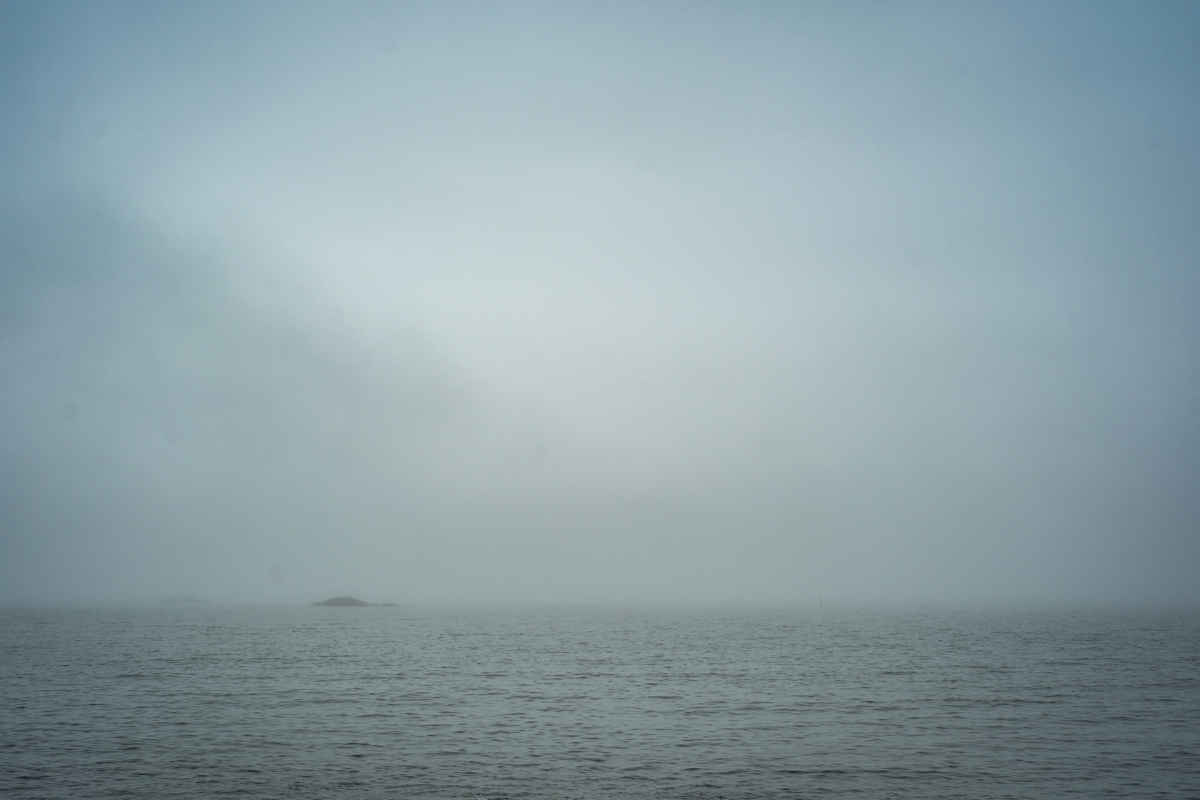 Misty seascape with calm gray waters under overcast sky, small distant island visible through fog, creating moody atmospheric scene