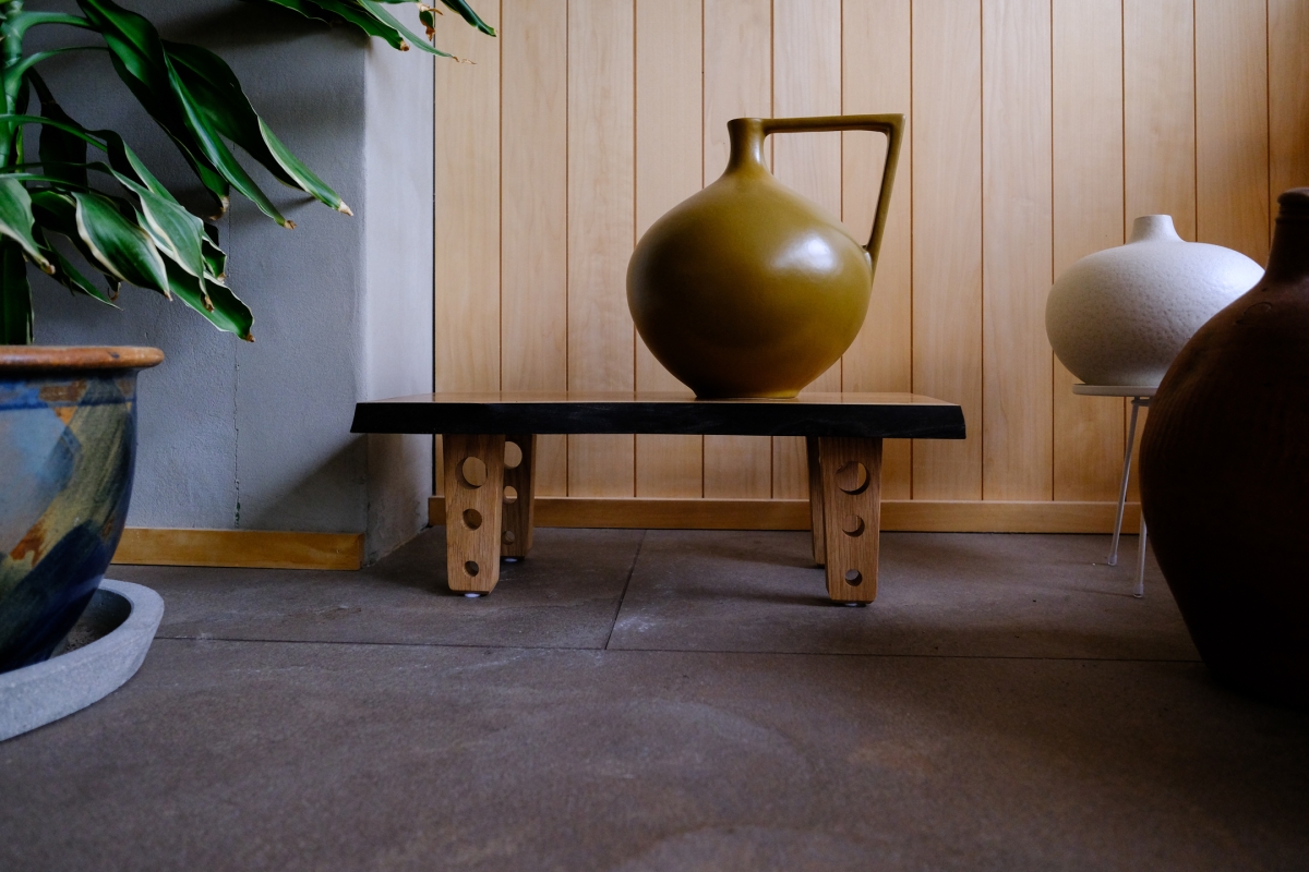 Modern wooden bench with decorative supports holding ceramic vases - olive green pitcher and white vessel against light wood paneling, with plant in foreground