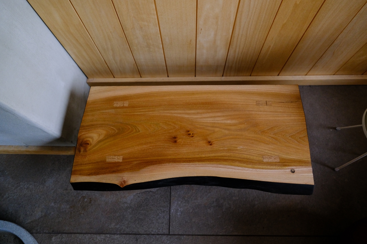 Live-edge wooden table with natural grain and knots positioned between white cushioned bench and dark floor tiles under wood-paneled ceiling