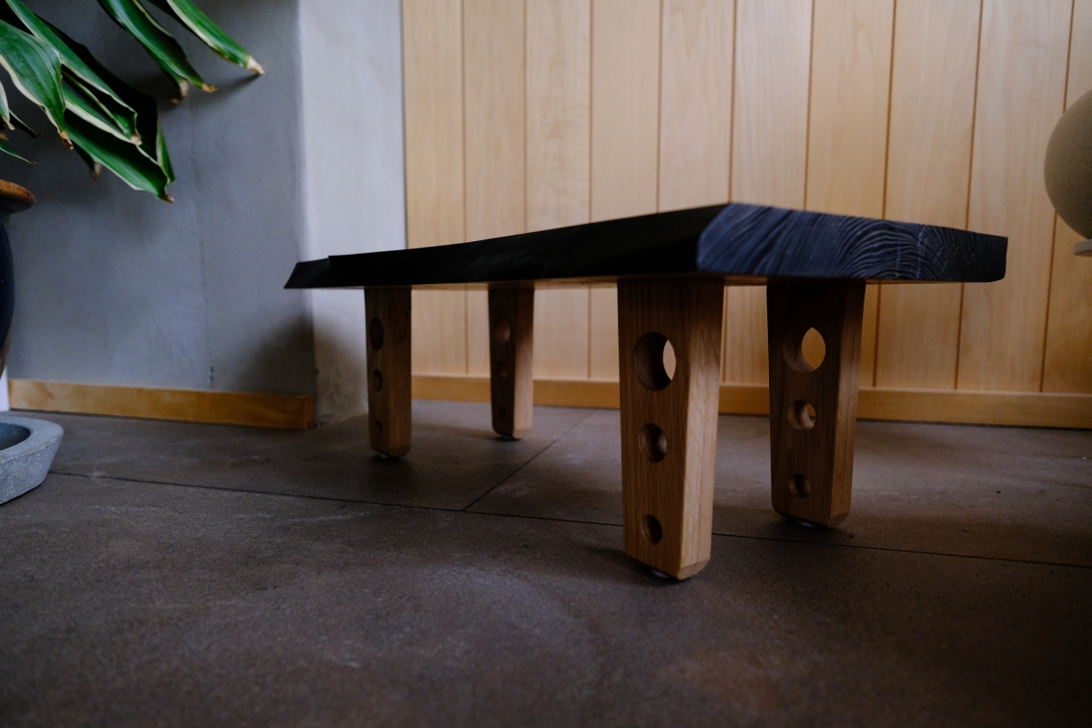 Handcrafted wooden bench with dark charred top and natural wood legs featuring decorative holes, positioned on dark floor against light wood paneling