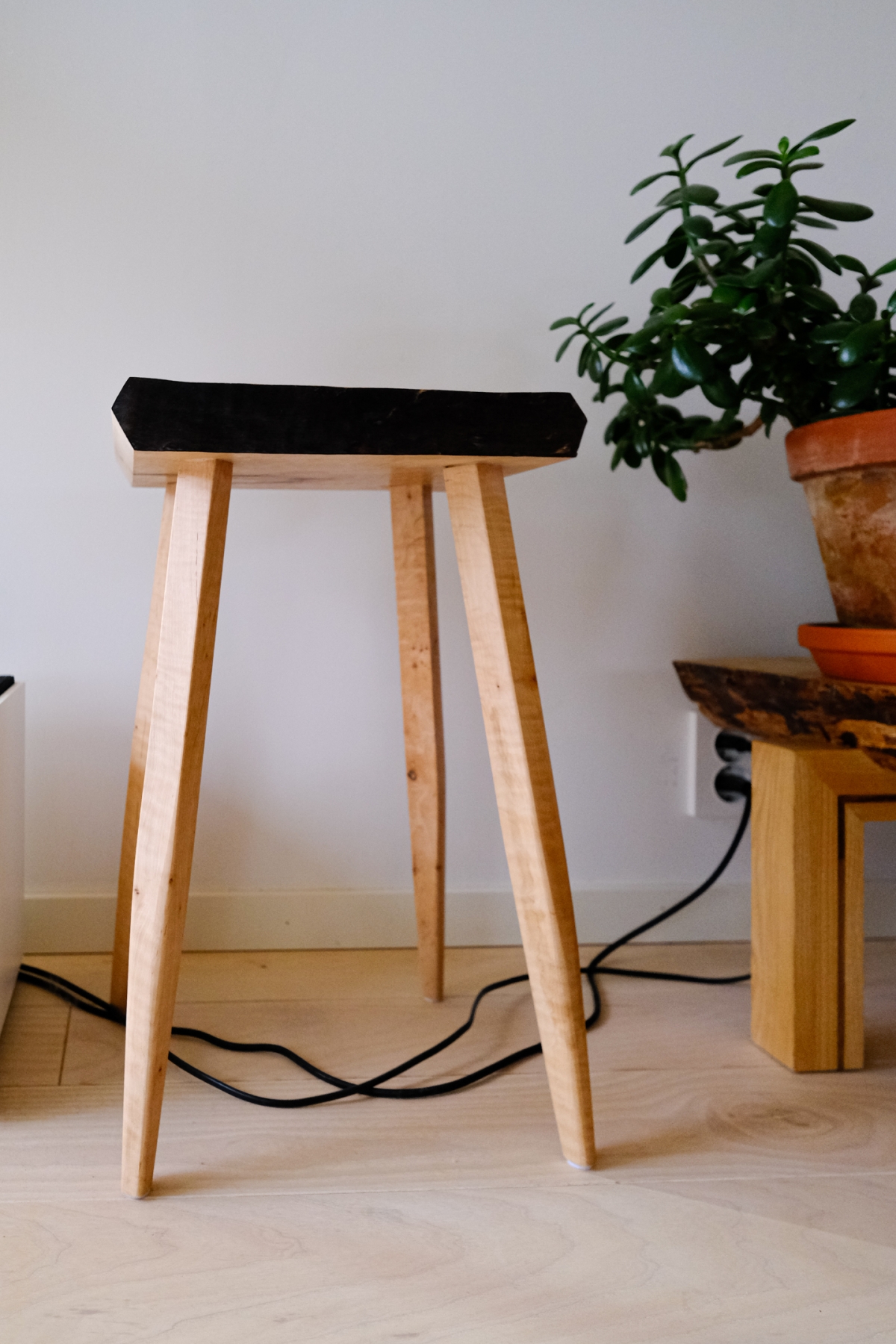 Minimalist wooden stool with black seat cushion and exposed electrical cables on light wooden floor, with potted plant on small table nearby