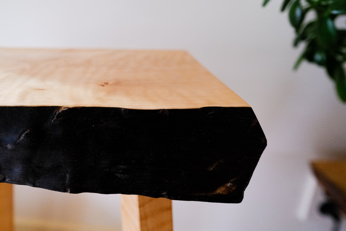 Live edge wood table corner with natural light wood top and charred black edge, showing organic grain patterns and rustic craftsmanship