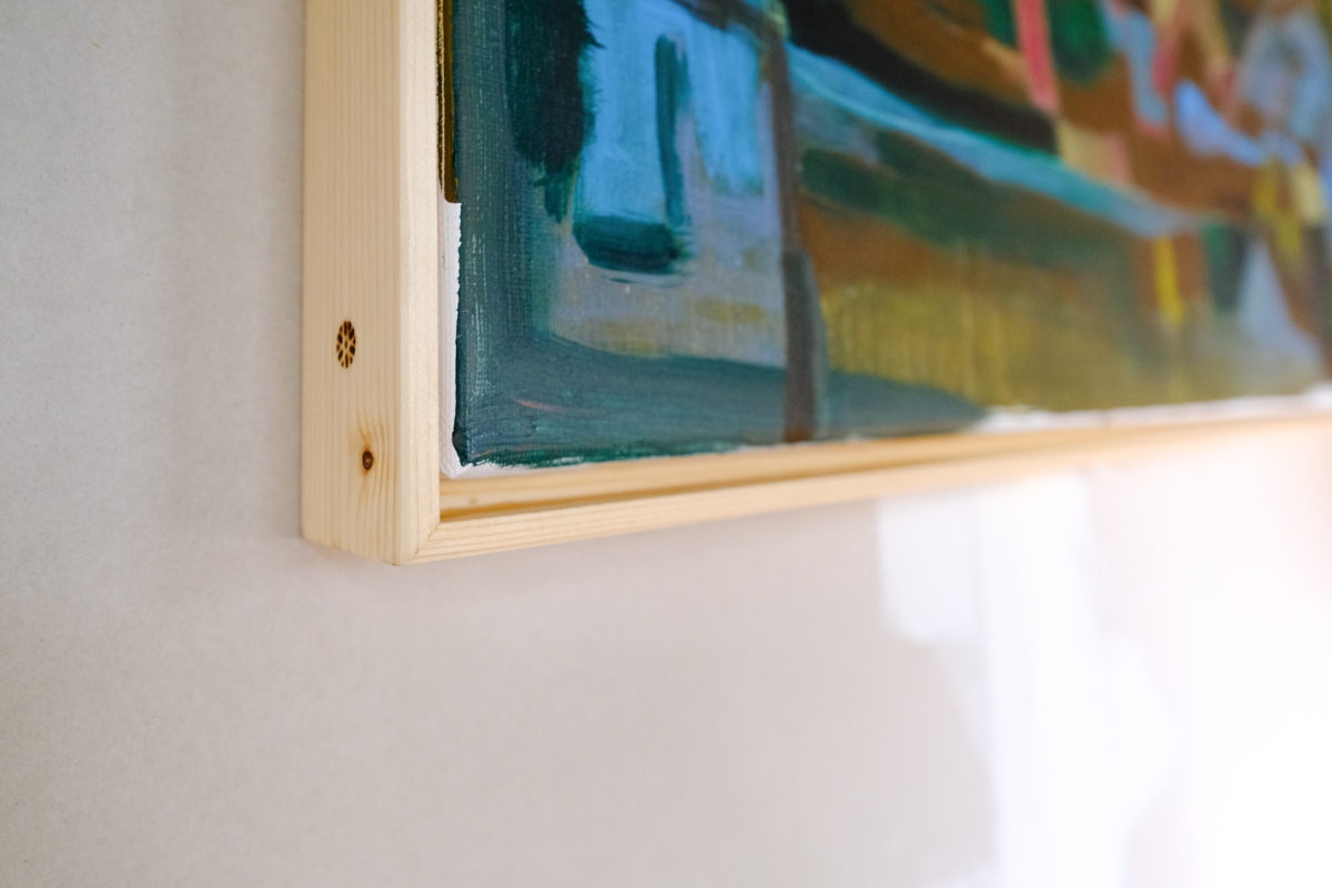 Corner of an unstretched painting on wooden stretcher bars, hung on a white wall, showing abstract blue, green, and gold brushstrokes.