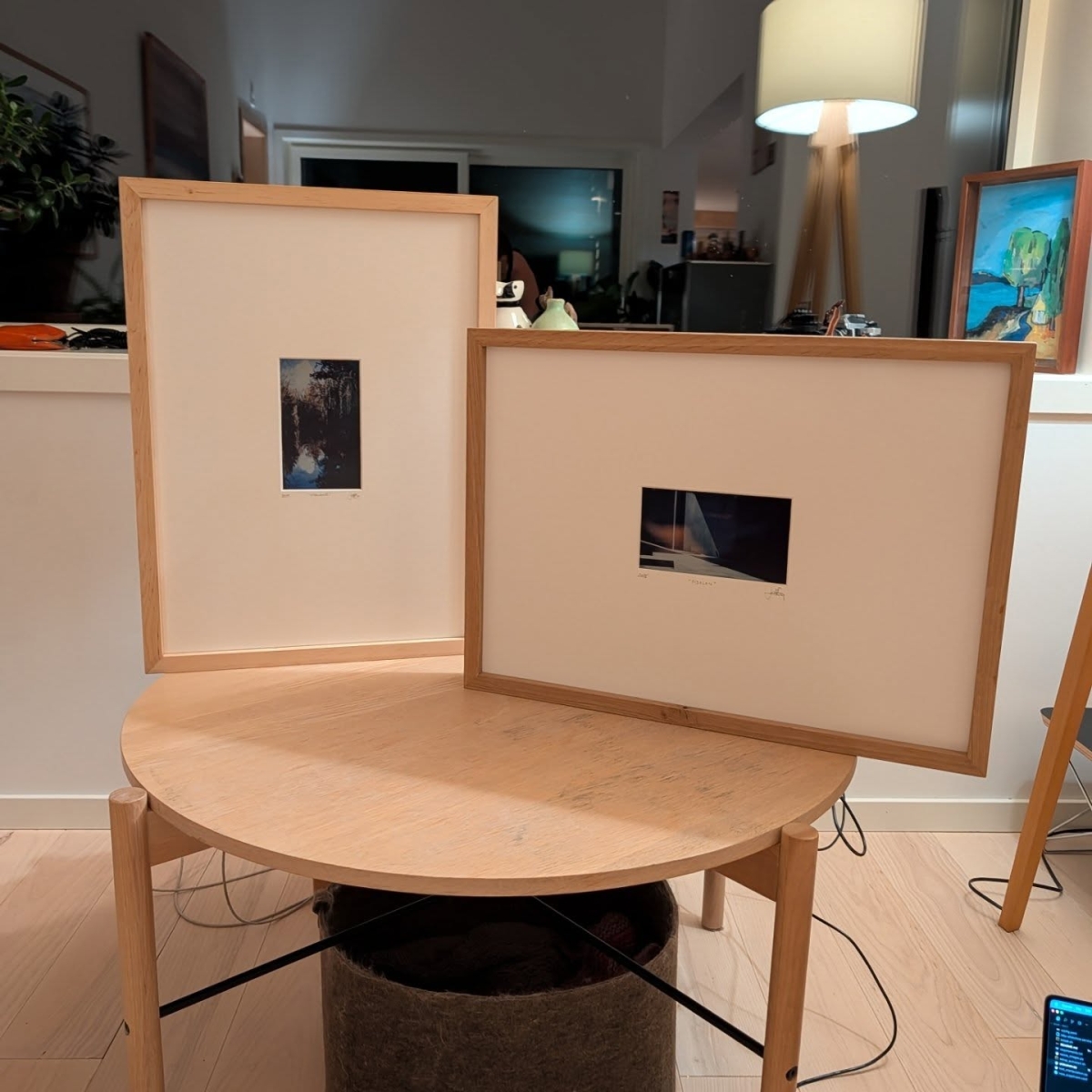 Two framed photographs with light wood frames displayed on a round wooden table in a modern living room with warm lighting