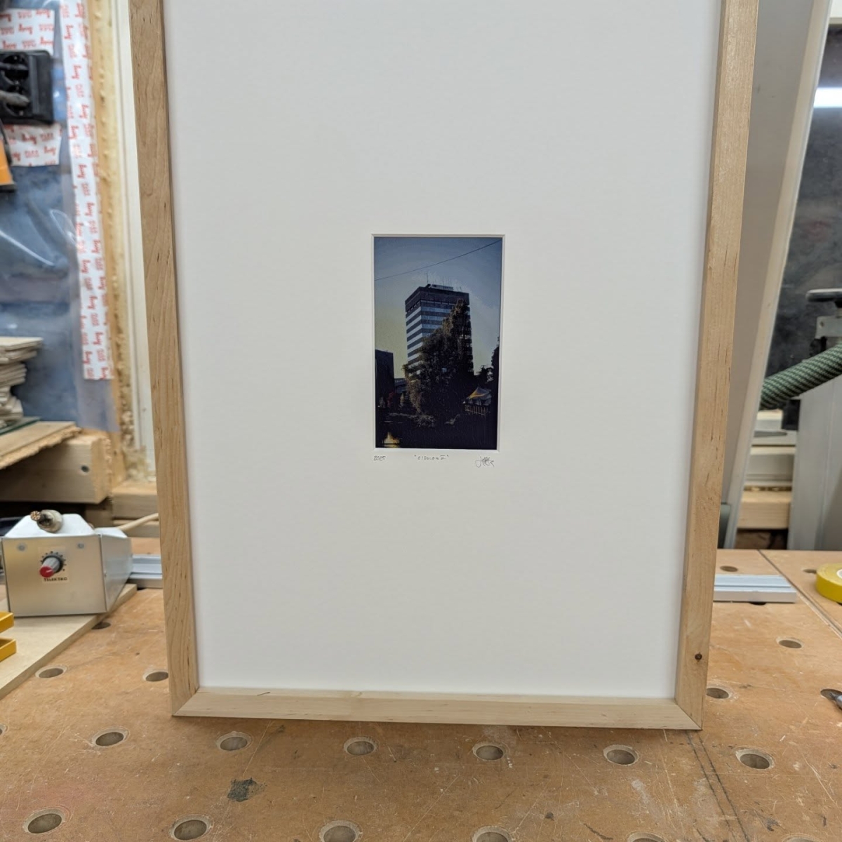 Framed photograph of a modern high-rise building at dusk mounted on white matting with light wood frame, displayed on a workshop bench