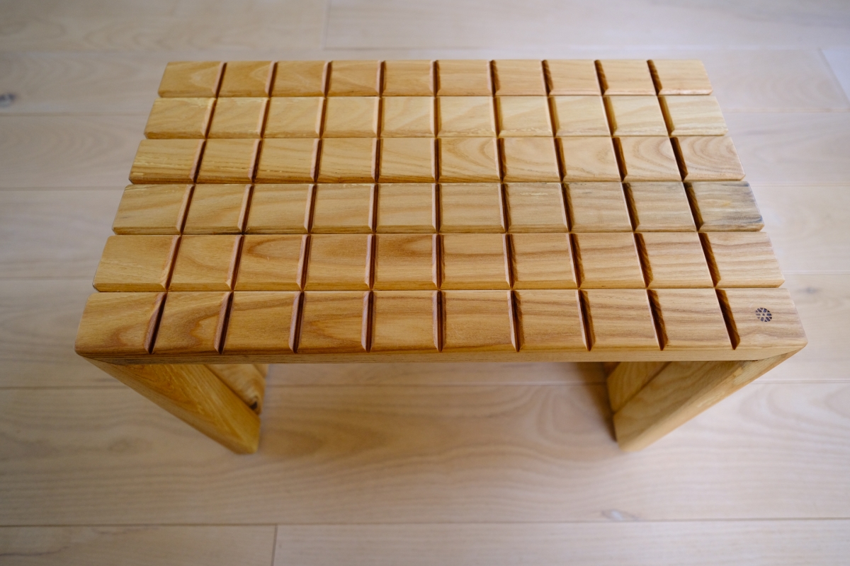 Wooden slatted stool with grid-like top pattern, X-shaped legs, natural light wood grain, set on pale wooden floor.