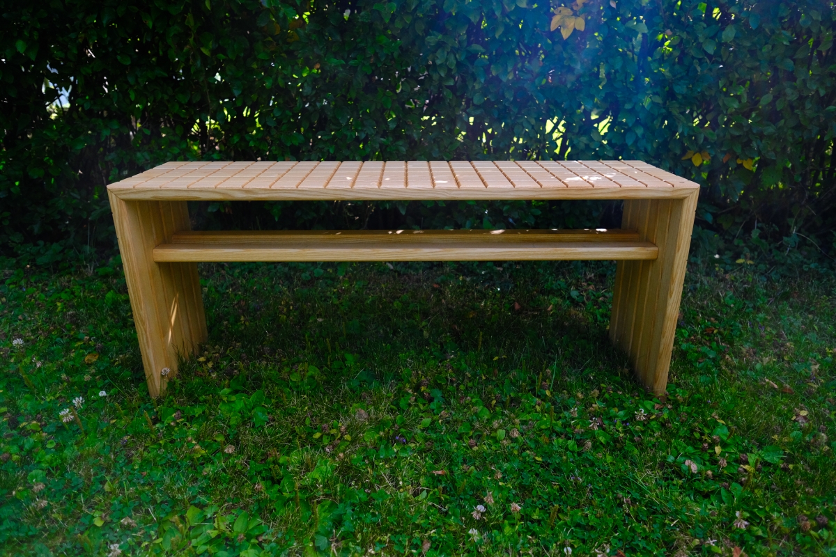 Wooden slatted bench with solid side panels sits on grass in front of dense green foliage, bathed in dappled sunlight.