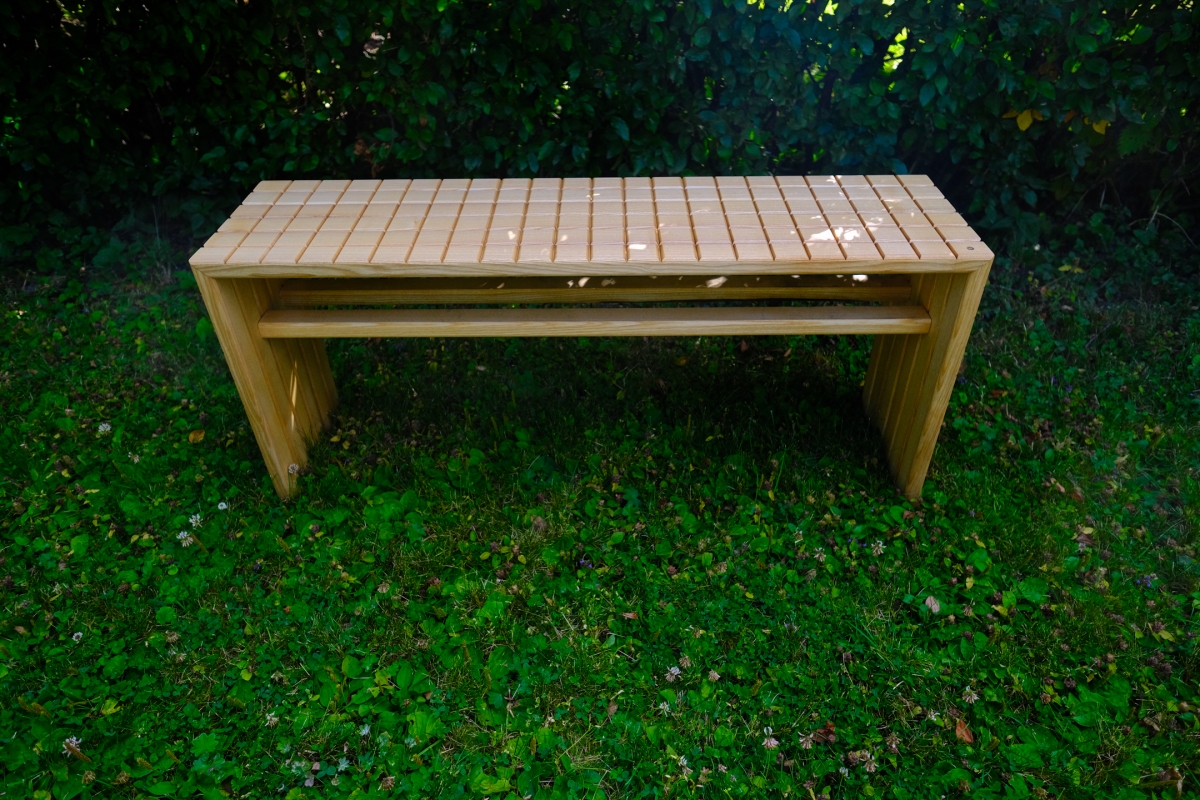 Wooden slatted bench with light natural finish, solid panel legs, set on a lush green lawn with clover and dense dark foliage behind.