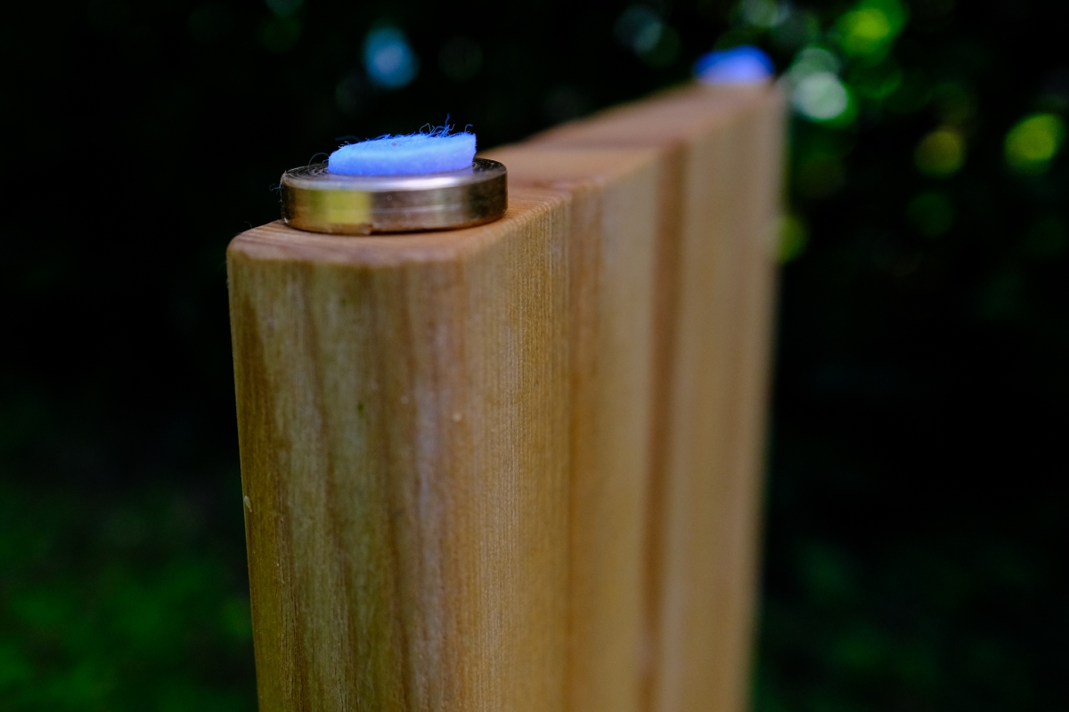 Close-up of a wooden post with a small round metal-rimmed object holding blue fibrous material, blurred green foliage in background.