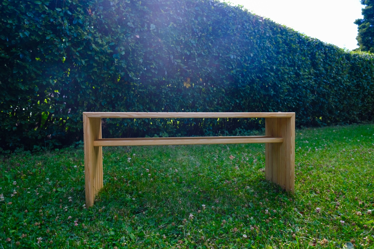 Wooden bench with solid panel legs and a round stretcher bar, sitting on a grassy lawn in front of a tall green hedge with sunlight.