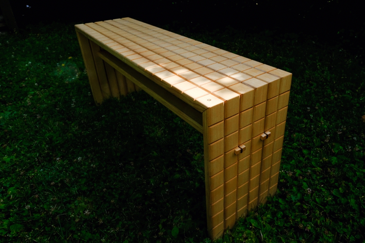 Wooden bench with a slatted, waffle-textured surface, sitting on dark green grass at night, illuminated by soft warm light.