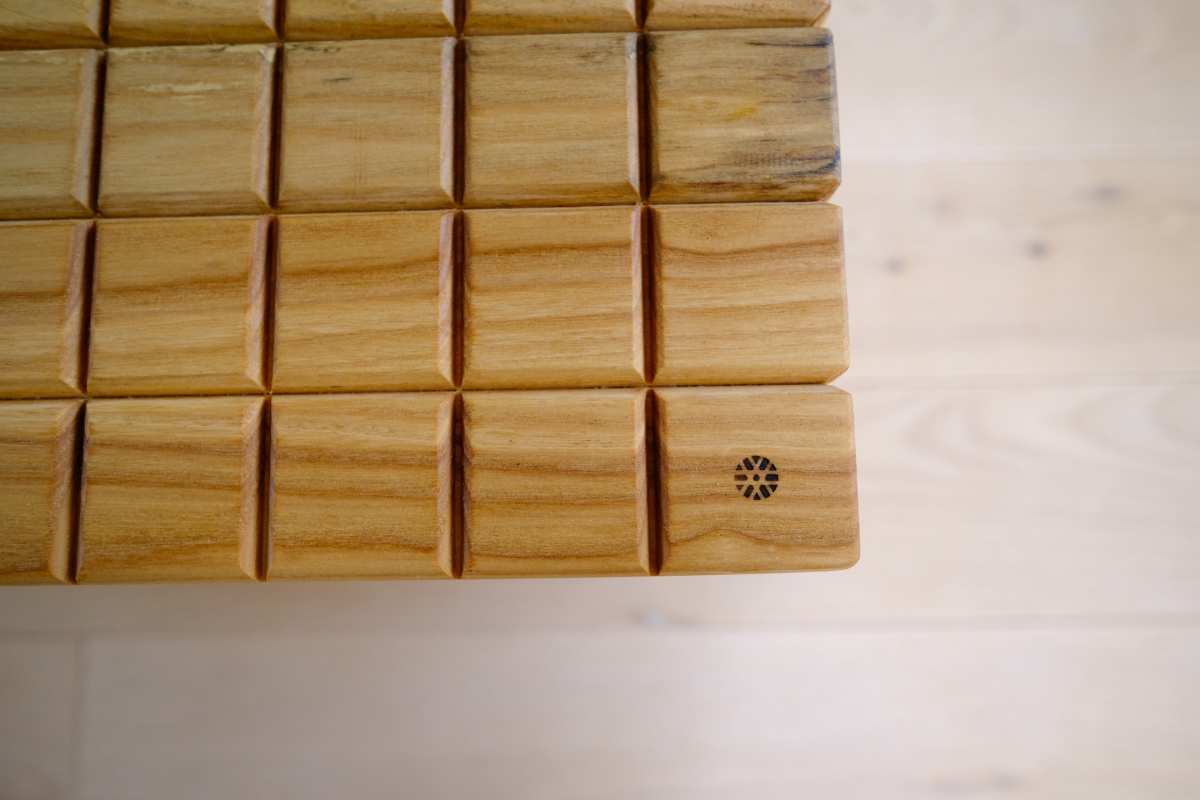 Wooden slatted surface with segmented blocks in a grid pattern, warm honey-toned wood grain, one block featuring a small compass-like engraving.