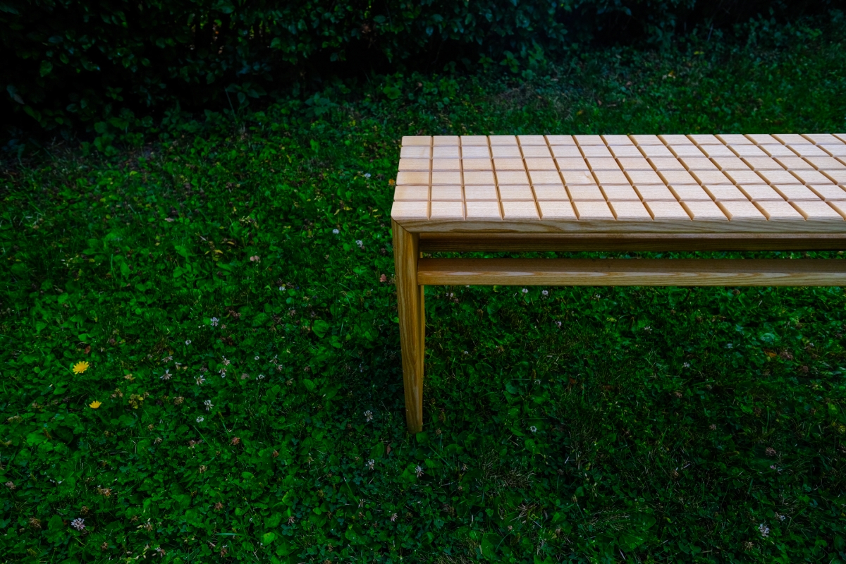Wooden bench with a woven lattice top sits on lush green grass dotted with clover and dandelions, set against a dark leafy backdrop.