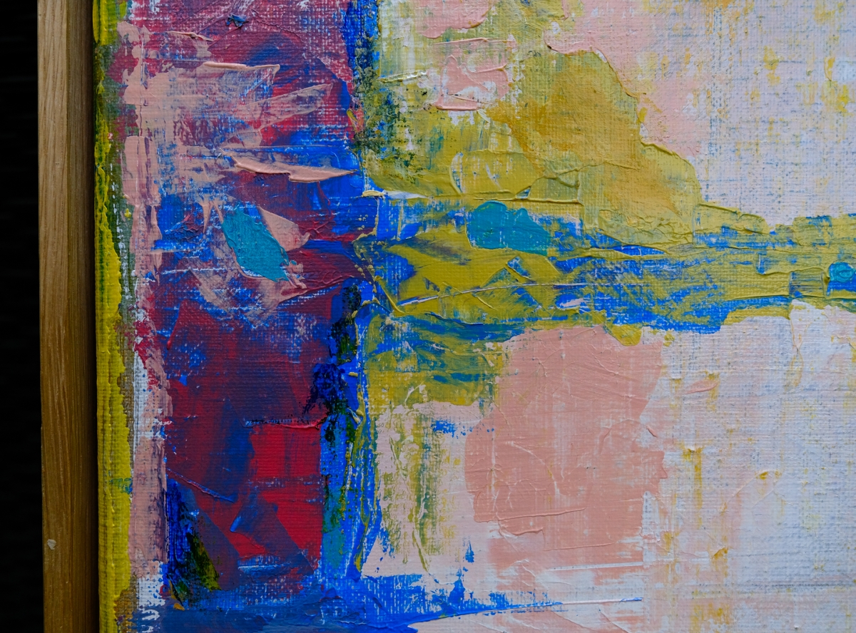 Close-up of an abstract painting with thick textured brushstrokes in deep red, blue, mustard yellow, and pink, edged by a wooden frame.
