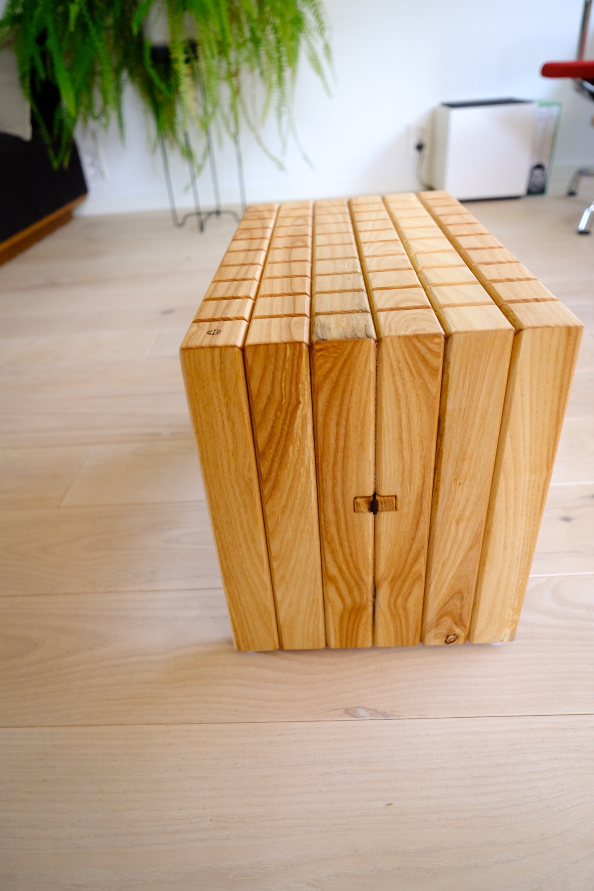 Wooden slat stool made of joined wood strips with visible grain, on a light hardwood floor beside a fern plant and white wall.