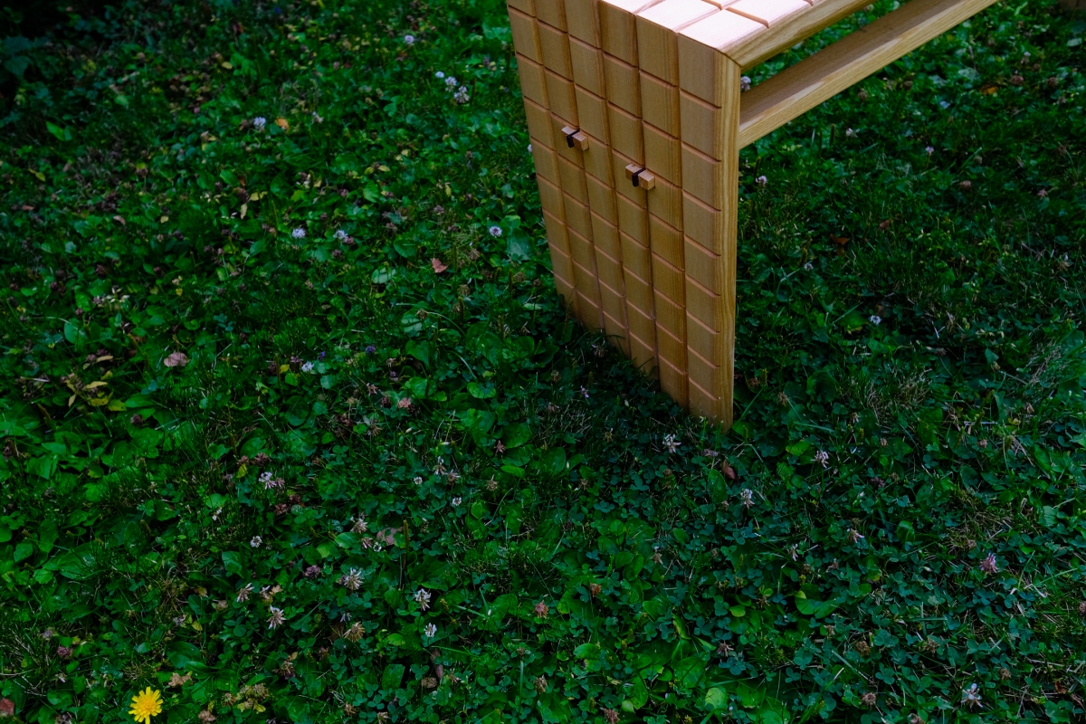 Wooden bench leg with slatted, grid-like design on lush green grass dotted with clover and small white flowers, viewed from above.