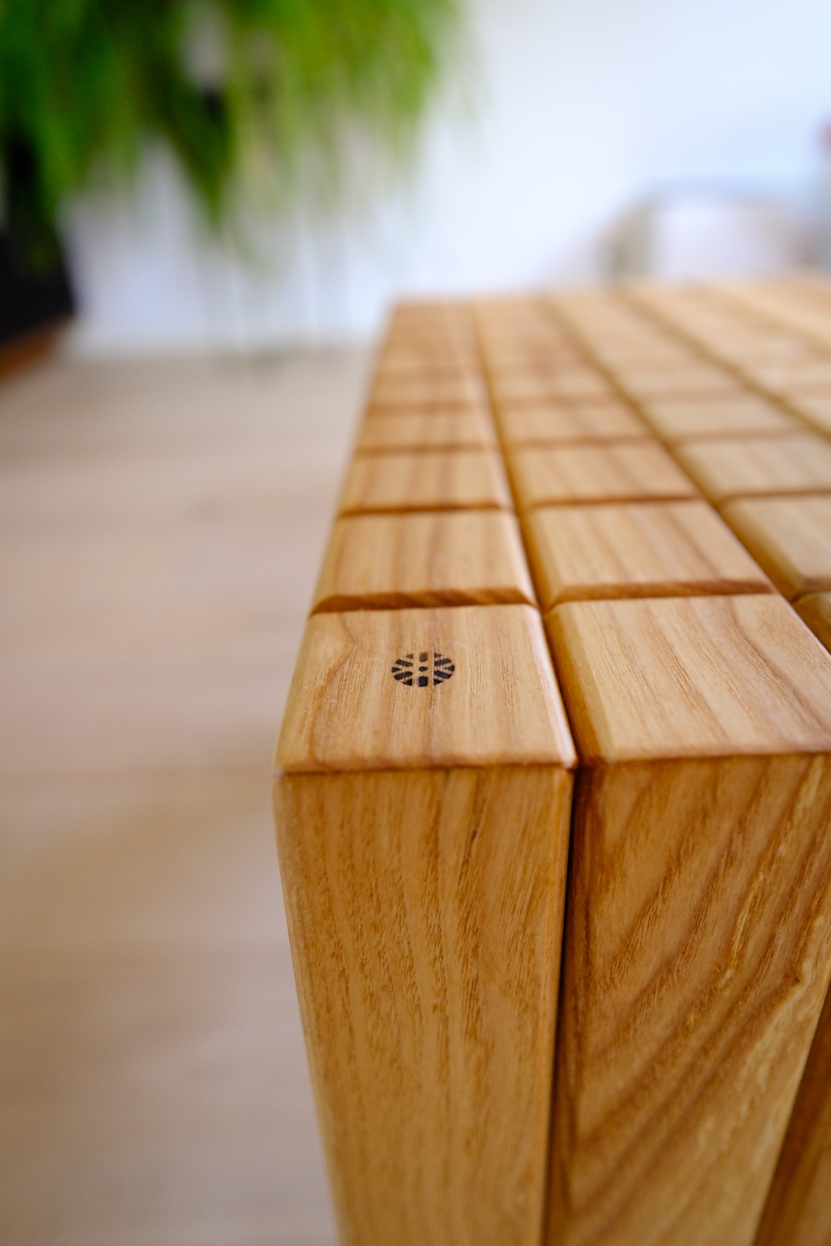 Close-up of a modern light wood table corner made from vertical wooden slats with a small circular emblem, blurred green plant in background.
