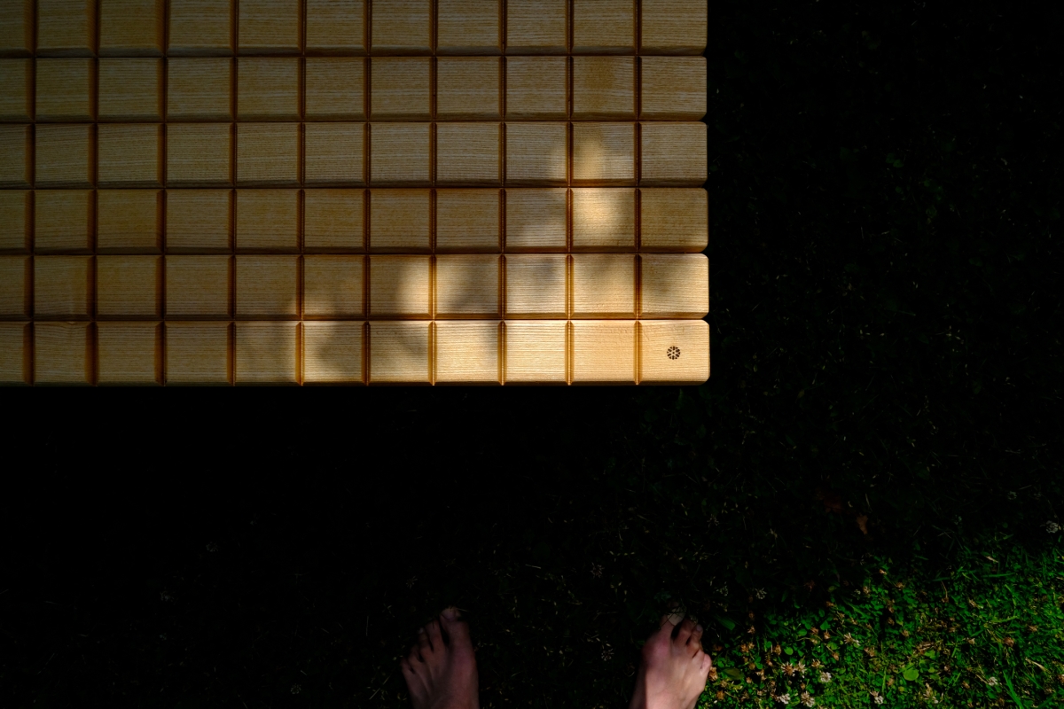 Overhead view of bare feet on dark grass beneath a sunlit wooden grid structure, tree shadow cast across its golden slats.