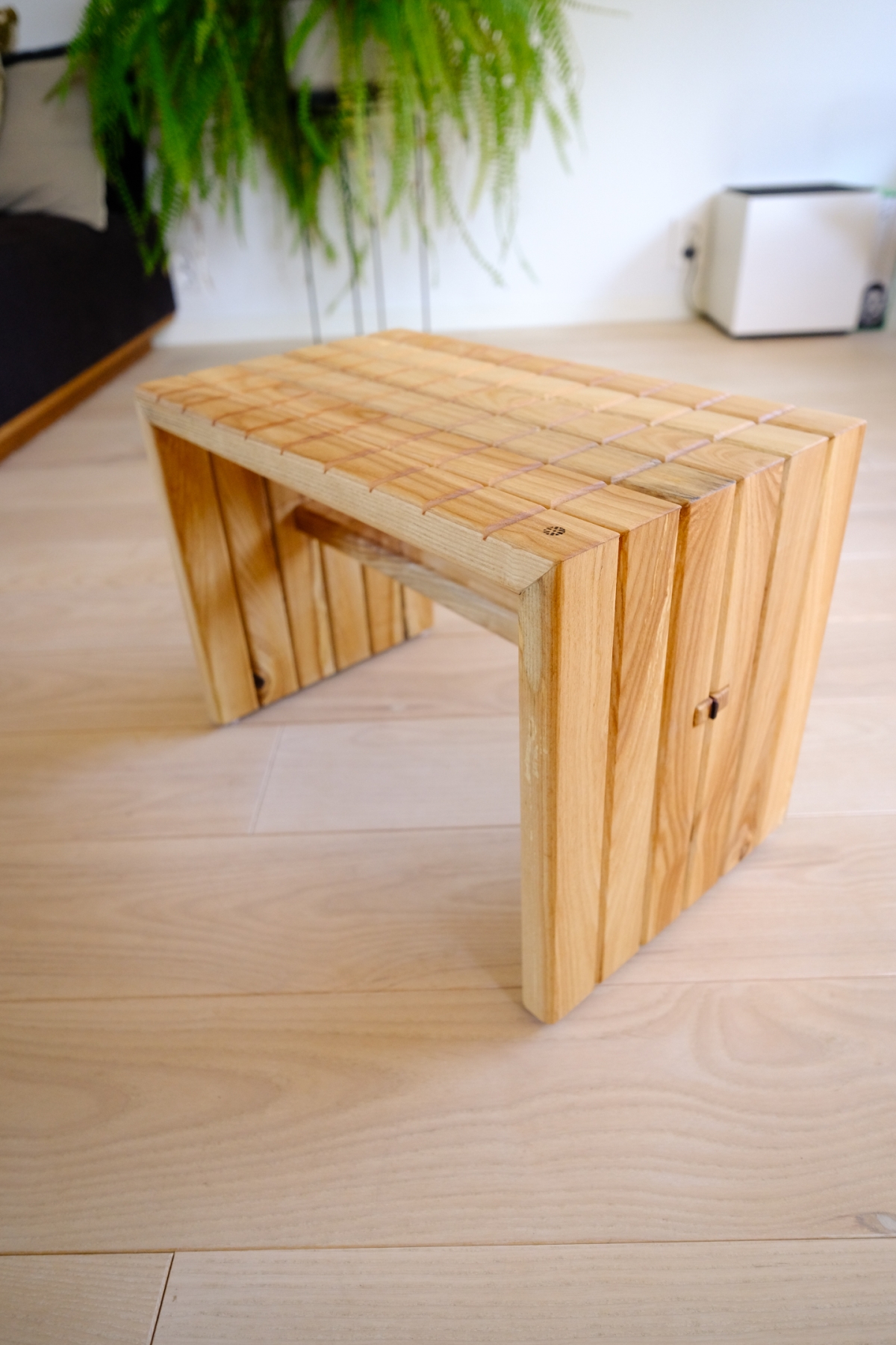 Handcrafted wooden slat bench with waterfall edge design, set on light hardwood floor near a hanging fern plant and white cabinet.