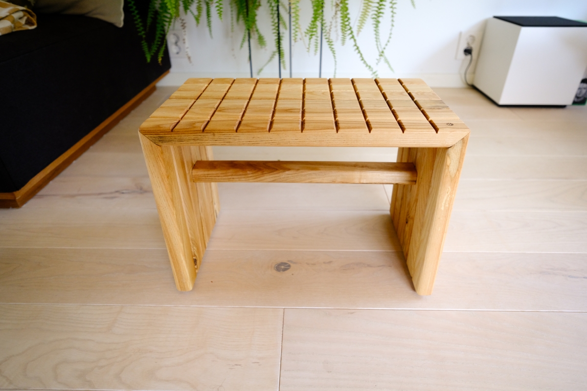 Small wooden slatted bench with panel legs and center support, on a light wood floor near a black sofa and hanging fern plant.