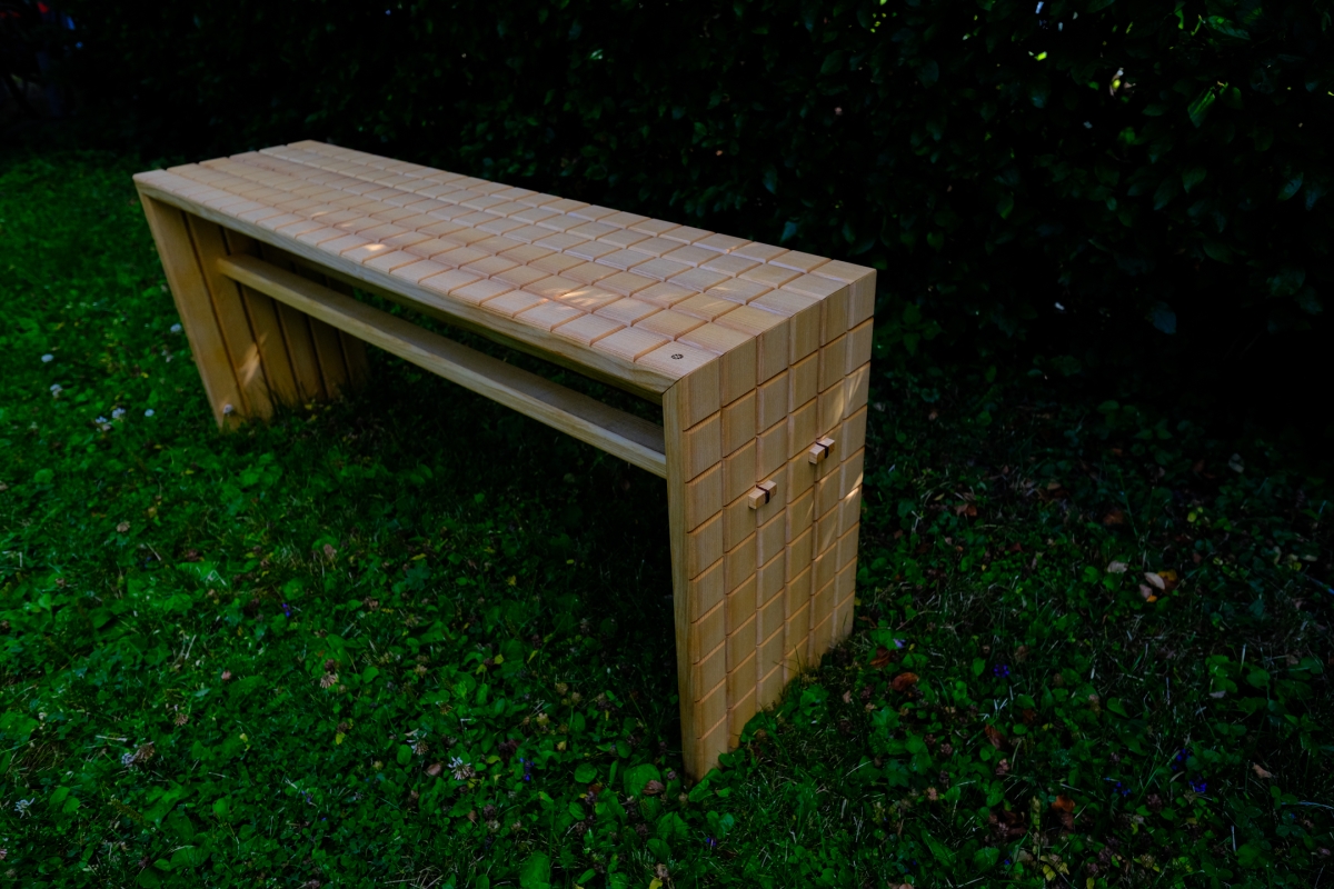 Wooden bench with tiled grid pattern and solid panel legs, standing on green grass against dark foliage backdrop at dusk.
