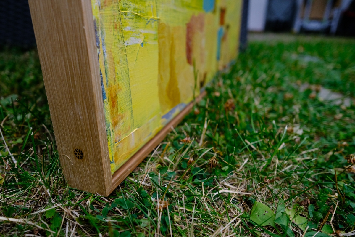 Close-up of a yellow abstract painting's wooden canvas edge resting on green grass, showing textured brushstrokes in yellow, blue, and rust tones.