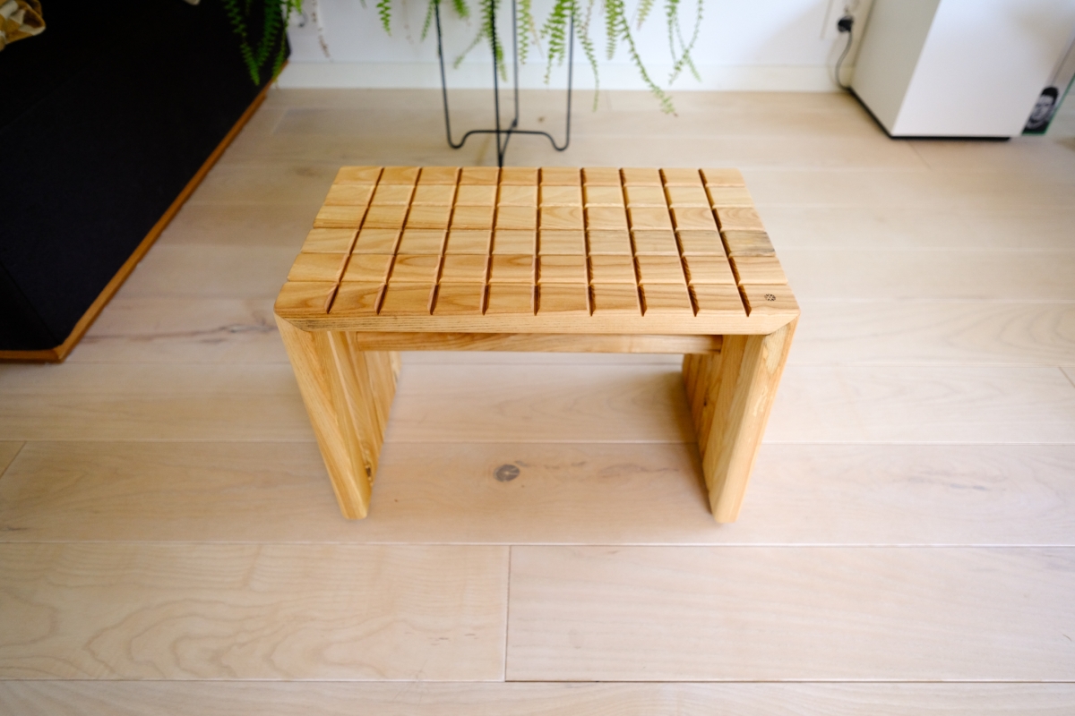 Small wooden stool with a grid-slat top and panel legs, sitting on a light hardwood floor near a plant stand and dark sofa.