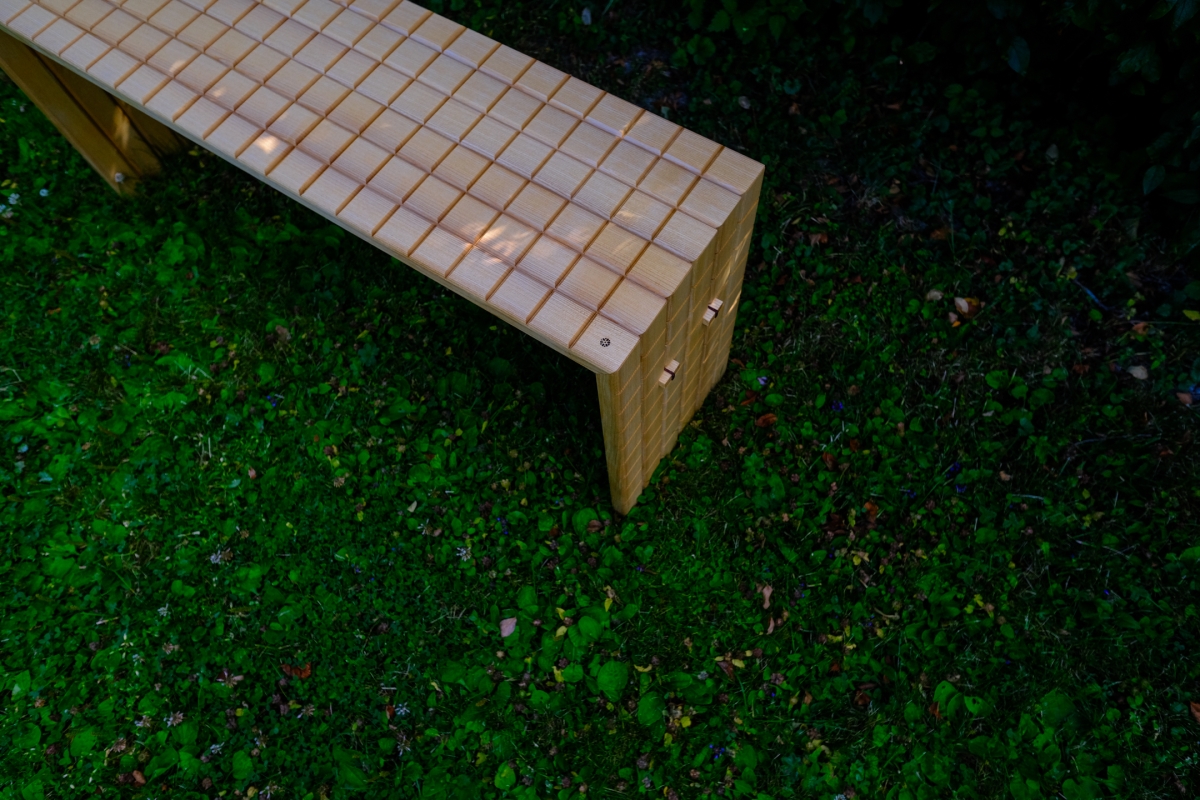 Overhead view of a wooden bench with a grid-patterned surface, angled diagonally across lush green mossy grass and scattered leaves.