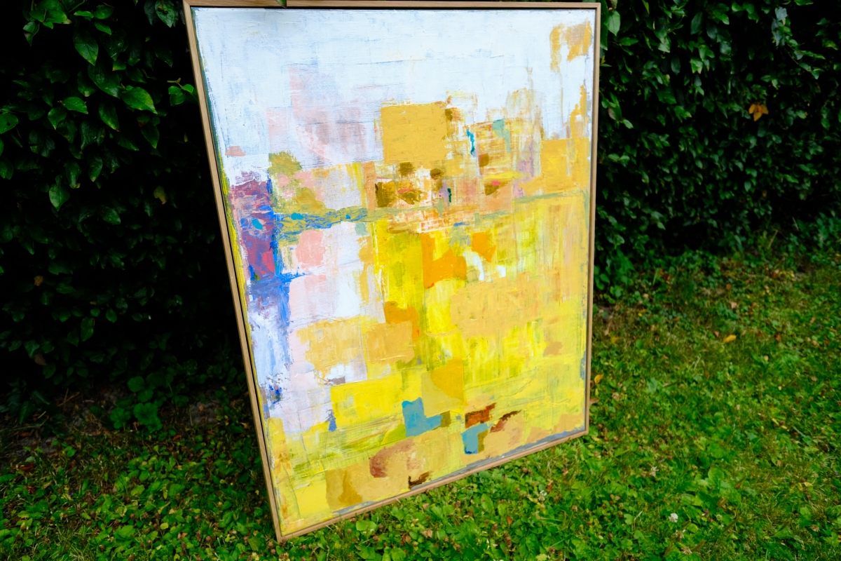 Abstract framed painting in yellow, gold, and white tones with blue and pink accents, leaning against a dark green hedge outdoors.