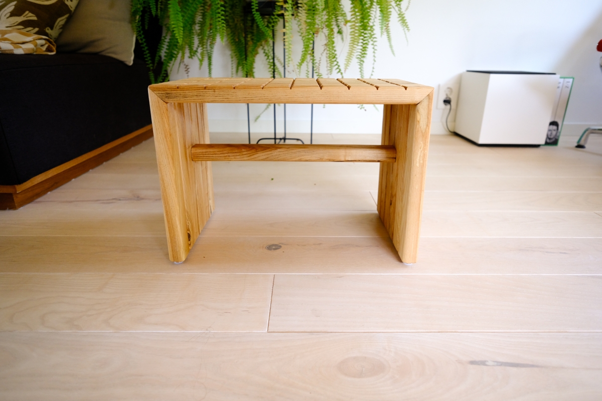 Wooden slatted stool with cross-brace support, sitting on light wood flooring beside a black sofa and hanging fern plant.
