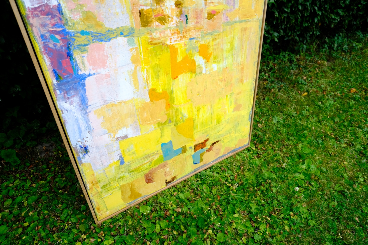 Abstract painting with yellow, blue, white, and maroon patches in a wooden frame, propped outdoors on green grass.