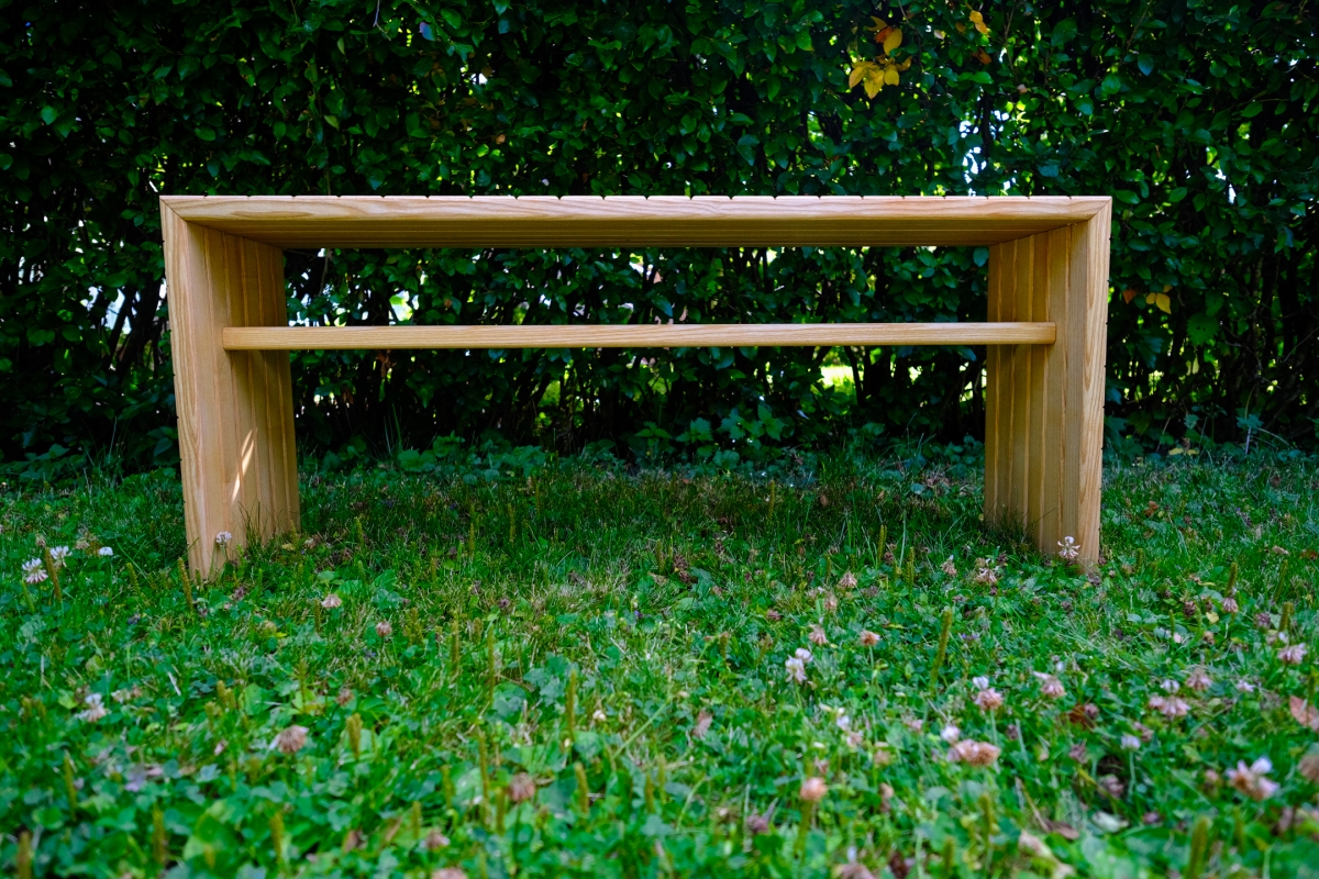 Wooden bench with slatted top and cross support, standing on a lush green lawn with clover, backed by dense dark foliage.