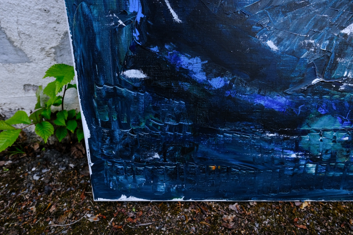 Abstract painting in deep blues and teals with thick brushstrokes leaning against white wall, green plant visible at left edge