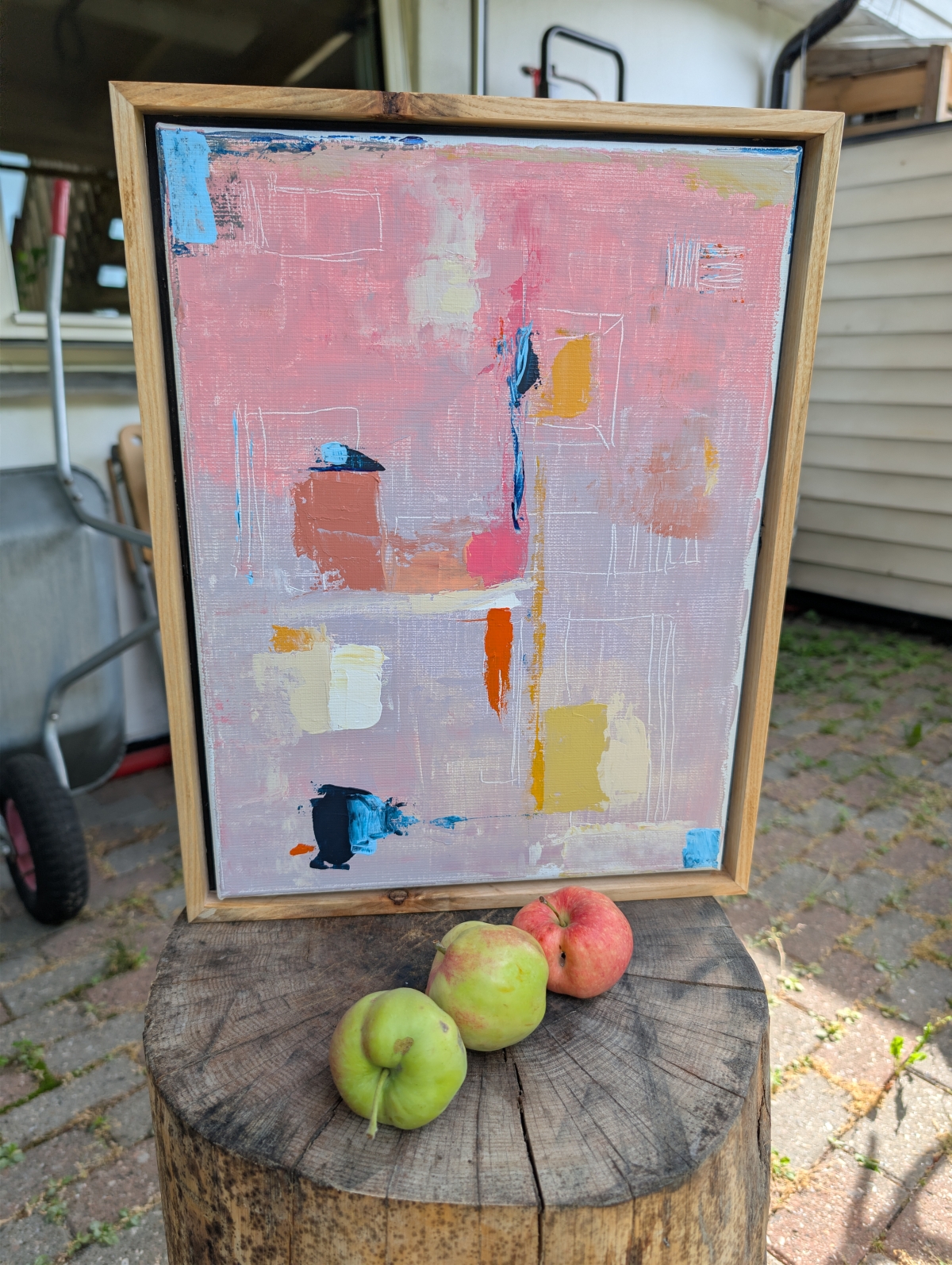 Framed abstract painting in pink, mauve, and gold tones on a wooden easel, resting on a tree stump with three apples beside it outdoors.