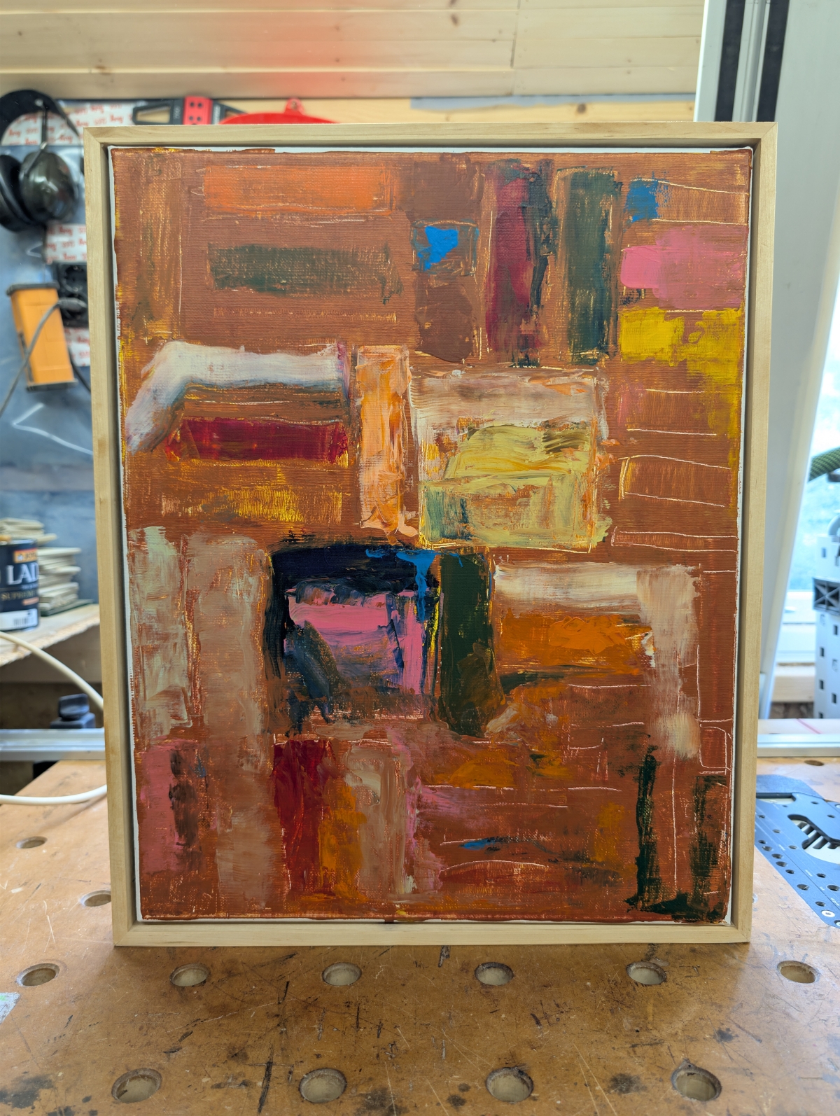 Abstract painting in a light wood frame, featuring rust-brown blocks with patches of blue, pink, yellow, teal, and orange, resting on a workbench.