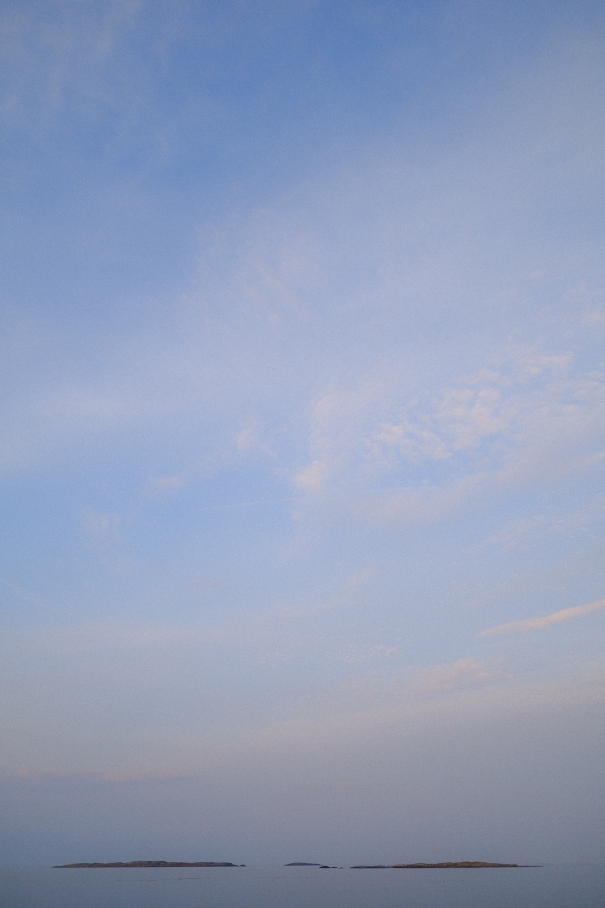 Vast pale blue sky with soft wispy clouds over calm water, small dark islets breaking the horizon line, tranquil minimalist seascape.