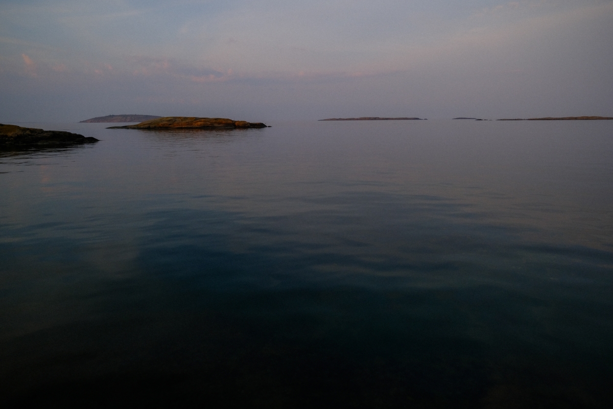 Calm sea at dusk with low rocky islets silhouetted on the horizon under a soft pink-grey sky, water darkening to near black in foreground.