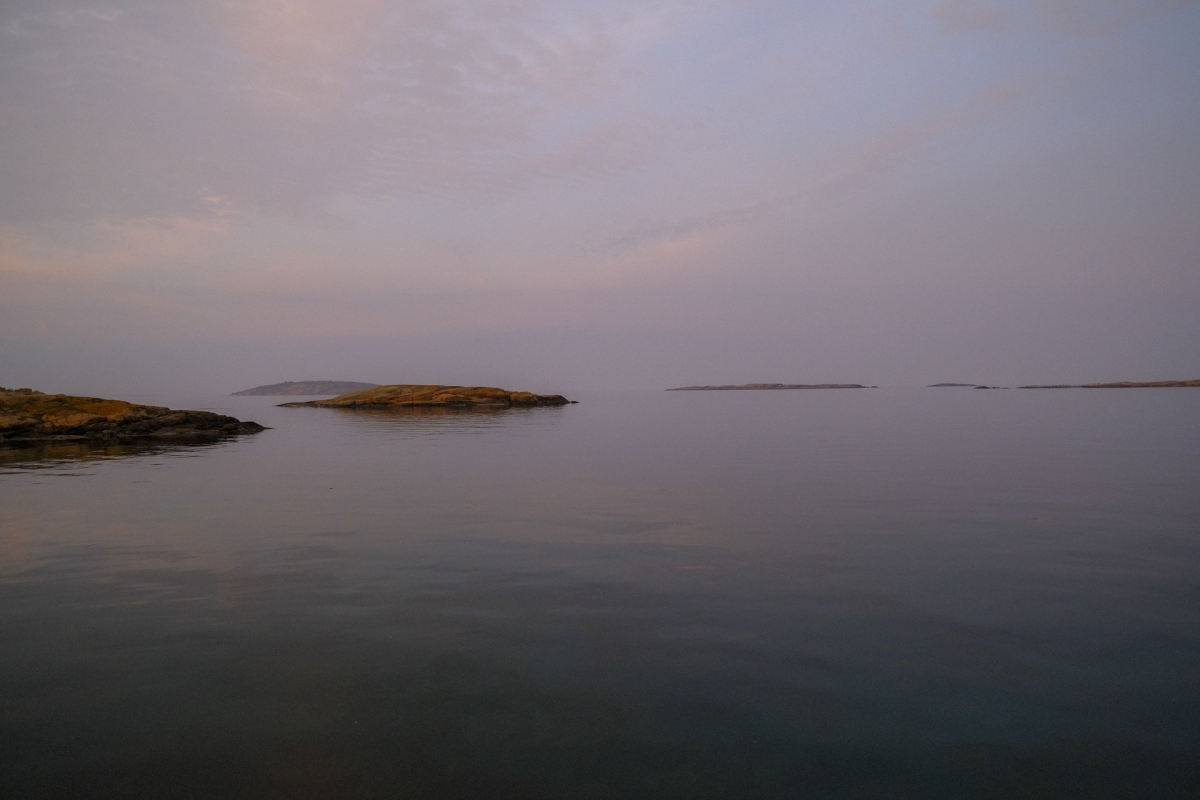 Calm sea at dusk with distant rocky islets, soft pastel sky in lavender and peach tones reflecting on still water.