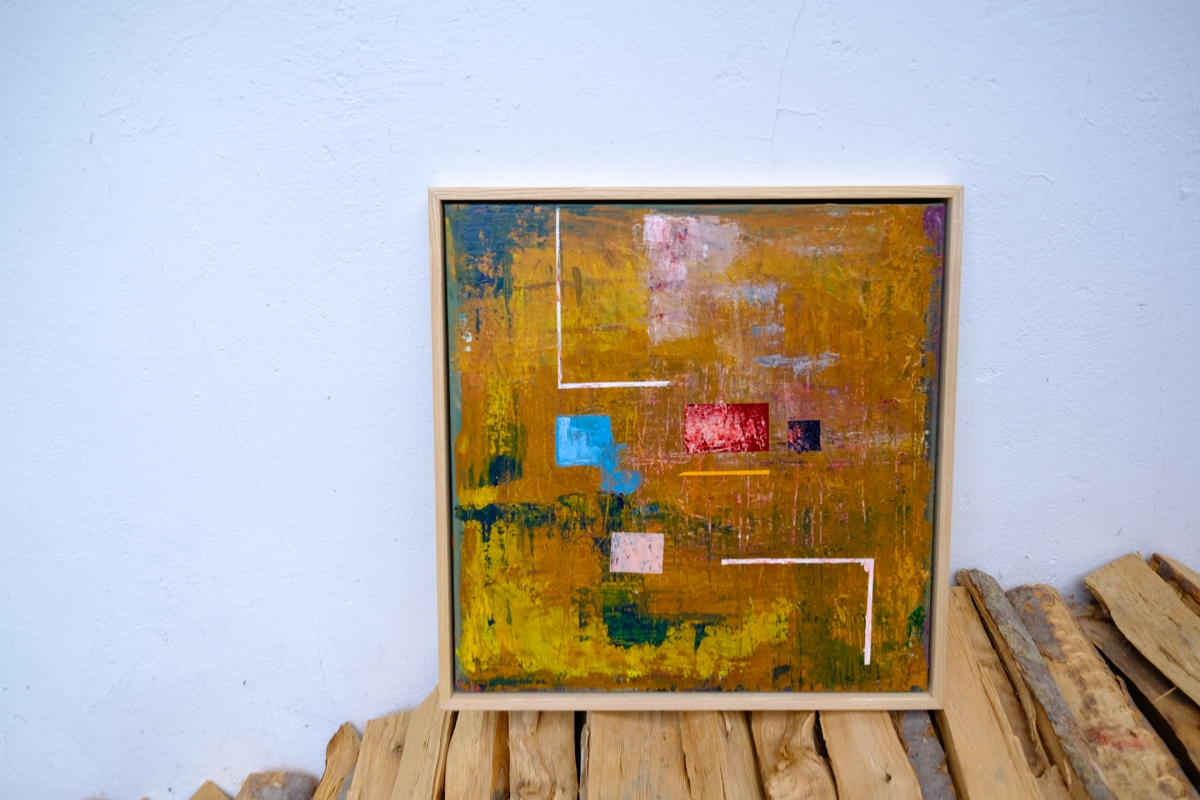 Abstract painting in a light wood frame with ochre background, blue, red, pink, and white geometric shapes, resting on stacked firewood against a white wall.