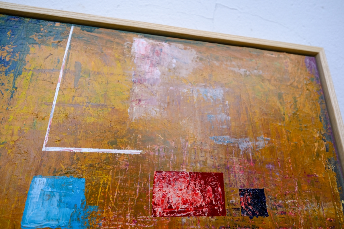 Abstract painting with textured golden-ochre background, geometric blue, red, and navy blocks, and a white L-shaped line, in a wooden frame.