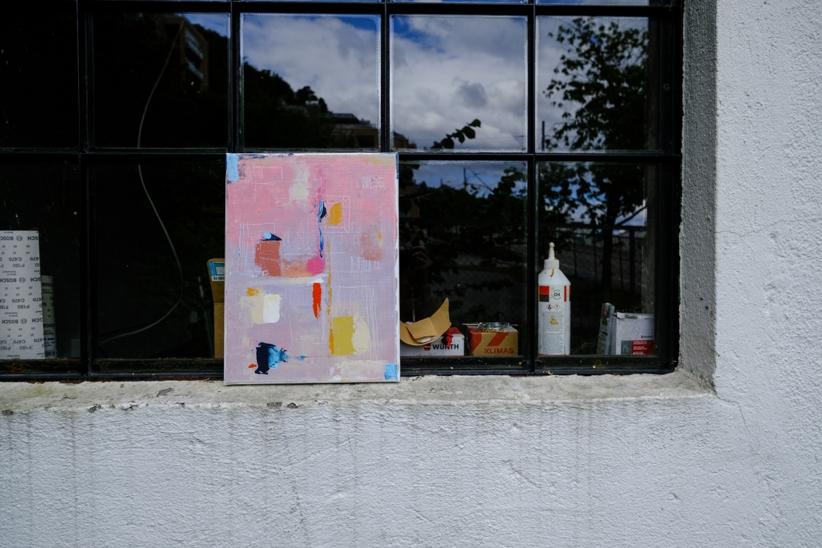 Abstract pink and pale purple painting with orange, blue, and cream accents, resting on a windowsill reflecting sky and trees.