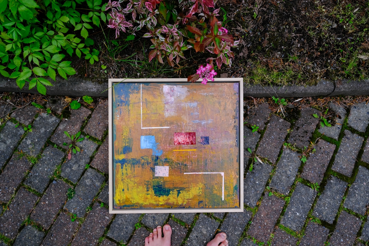 Framed abstract painting in gold, blue, and red tones lies on a brick path near green foliage; bare feet visible at bottom edge.
