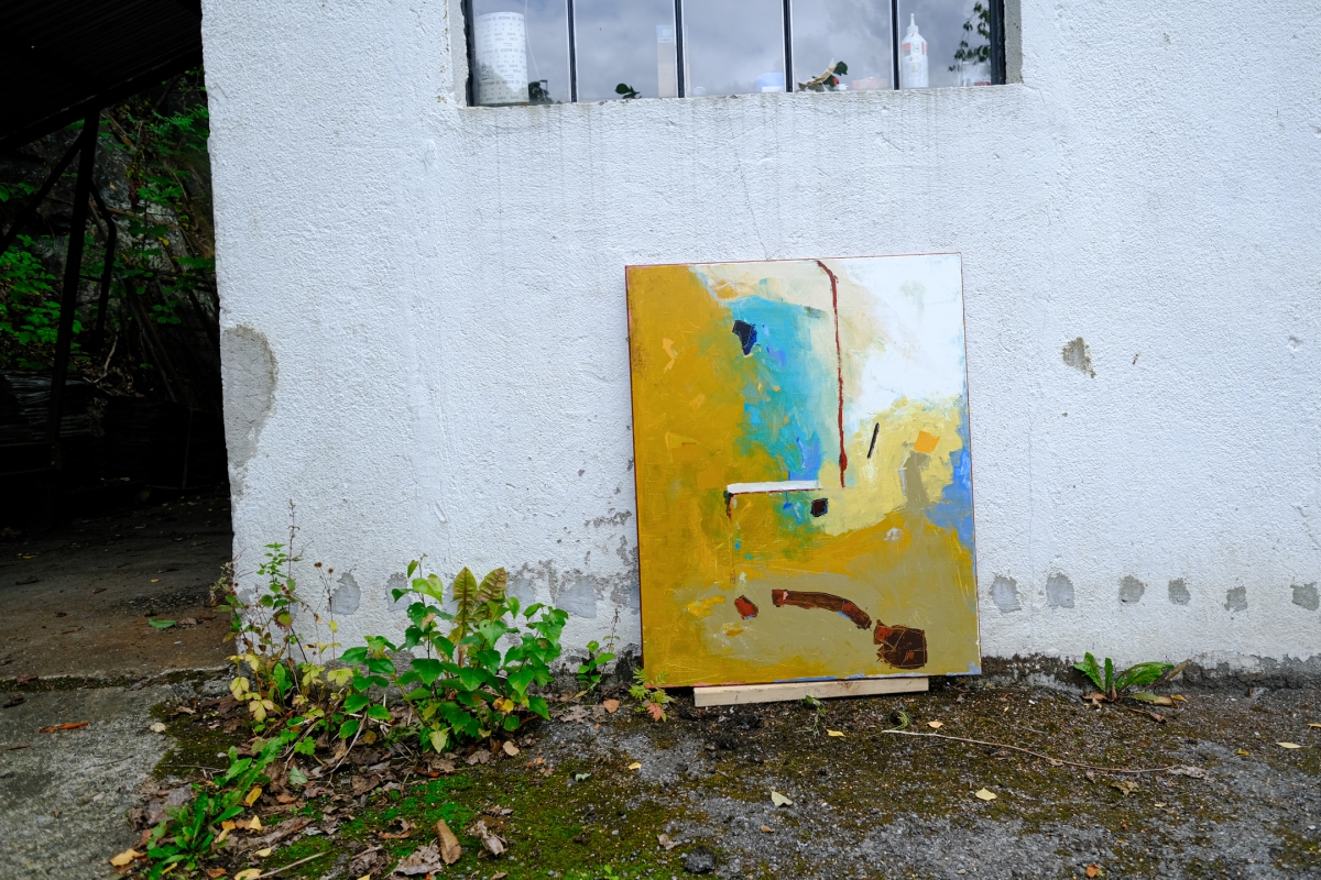 Abstract painting in ochre, teal, and cream tones leaning against a whitewashed wall, propped outdoors near weeds and a barred window.